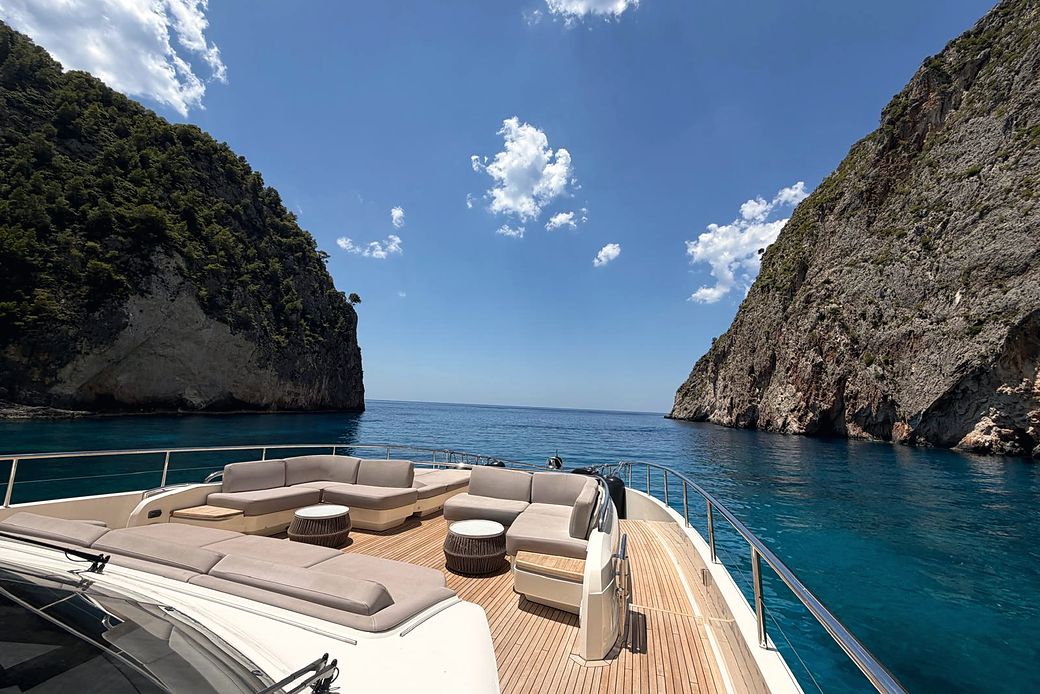 Yacht foredeck with cushioned seating and teak tables, at anchor in a turquoise cove between cliffs