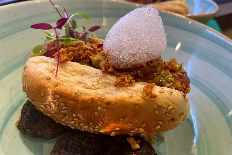 Sesame bun filled with tartare and avocado, with crispy onion, micro herbs and a foam, on a turquoise plate