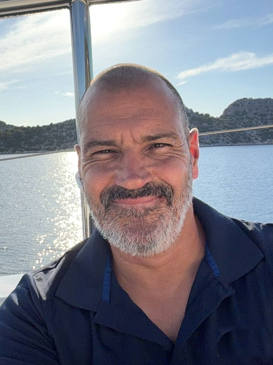 Jose Manuel Pereira smiling on deck in his crew polo, sunlit water and a rocky coast behind him